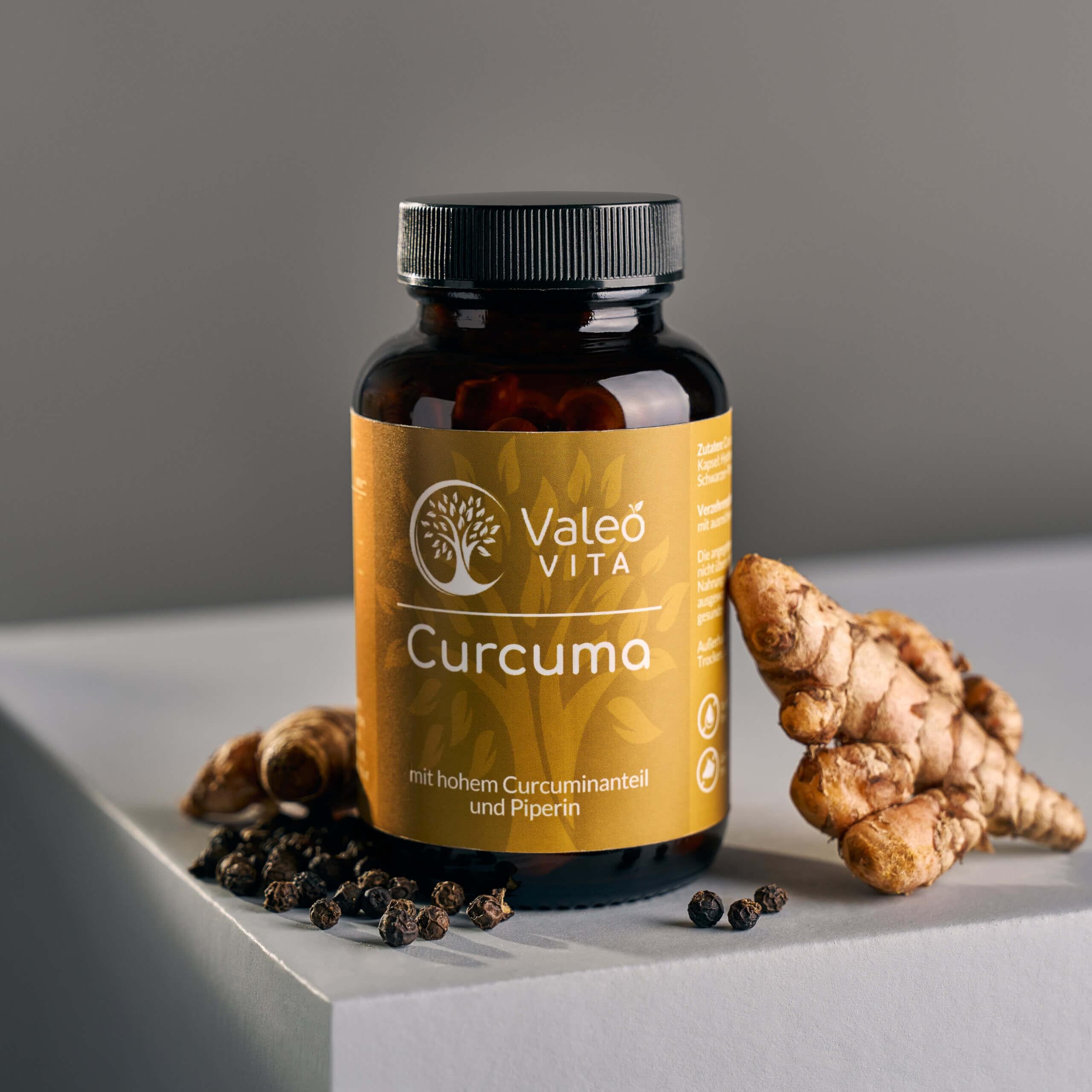 Valeo Vita Curcuma Kapseln in Braunglasdose mit Kurkumawurzel und schwarzen Pfefferkörnern auf hellem Podest