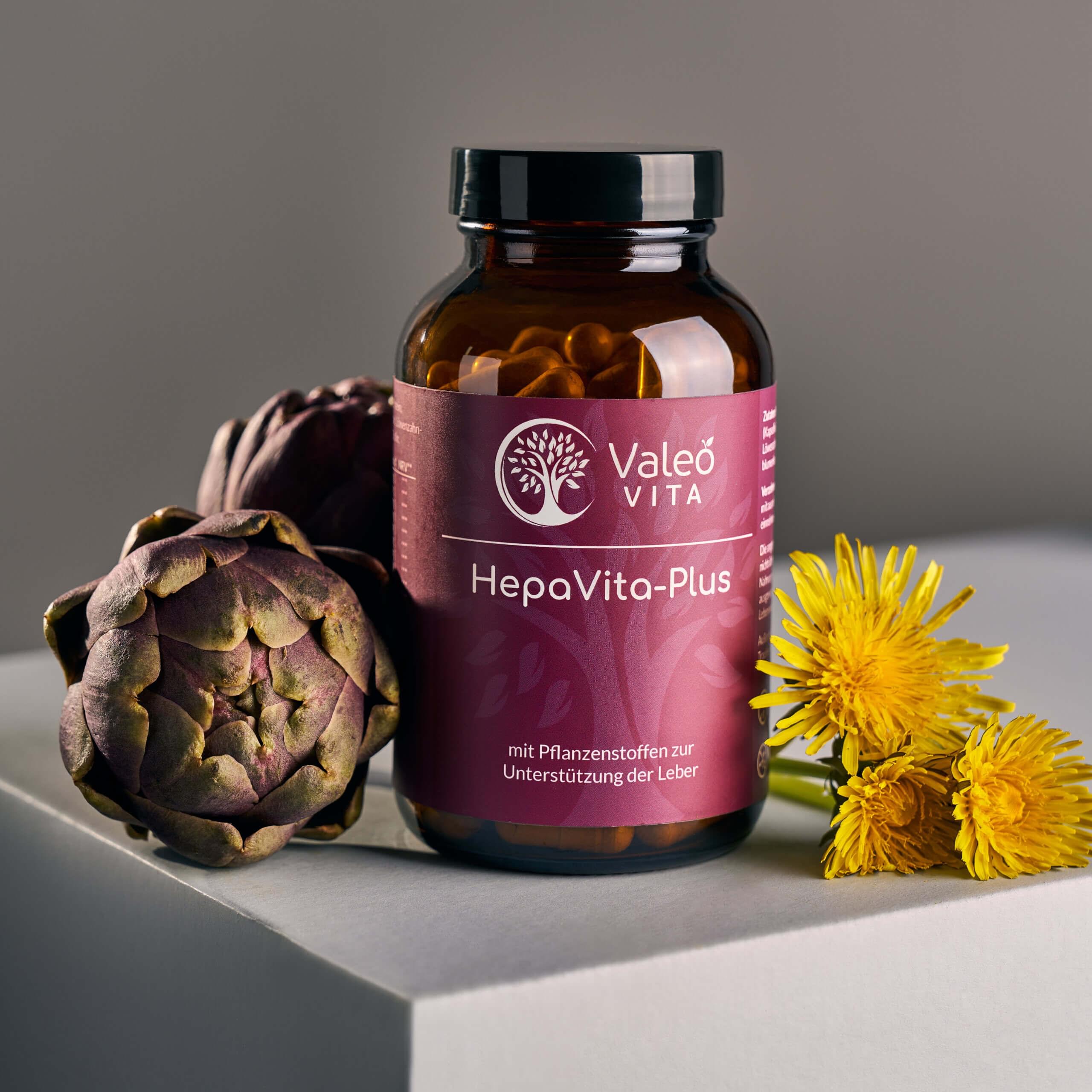 Valeo Vita HepaVita Plus Kapseln in Glasdose mit Artischocke und Löwenzahnblüten auf hellem Podest